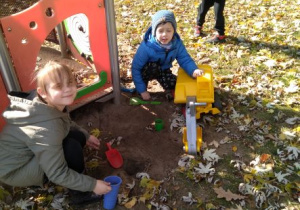 Dziewczynka i chłopiec bawią się koparką i przyborami do zabawy w piaskownicy. Tuż za nimi stoi drugi chłopiec